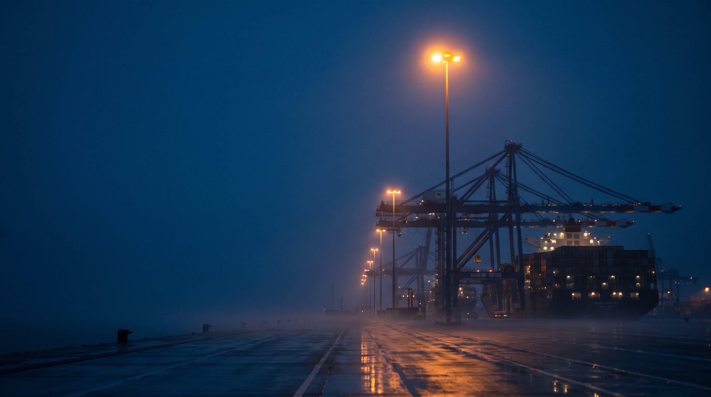 Terminal à conteneurs à l’heure bleue : quai désert sous une file de lampadaires ambrés, portiques et porte-conteneurs à droite